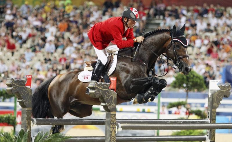 Le cavalier Eric Lamaze en veste rouge et son petit étalon bai Hickstead en pleine trajectoire de saut au-dessus d'un oxer géant décoré de dragons chinois aux Jeux Olympiques de 2008